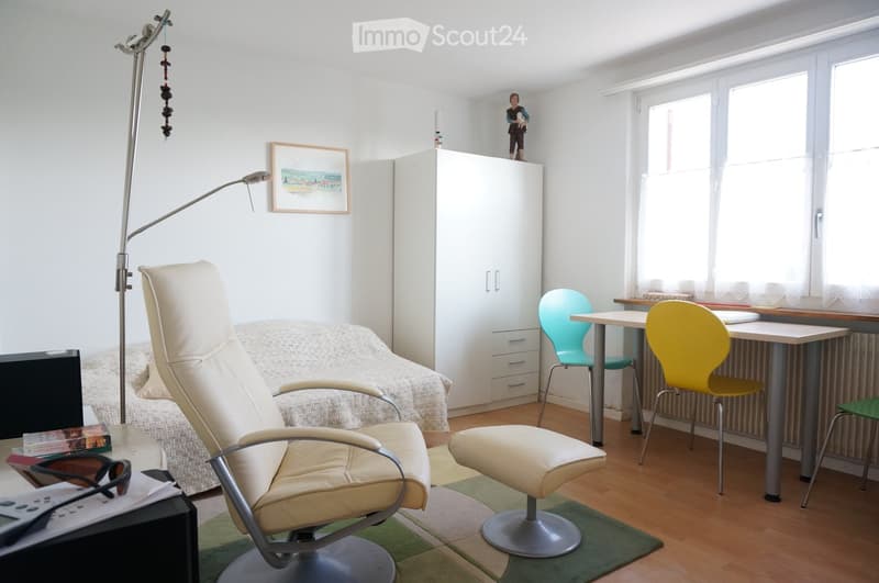A well-lit room with a sofa, a chair, a small table, a stool, a cabinet, a picture frame, and a floor lamp. The room has wooden flooring and large windows.