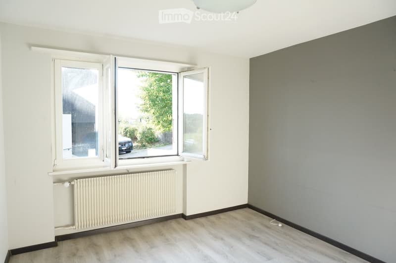 Empty room with light-colored walls, wooden flooring, and large windows with open shutters, allowing natural light to enter. A radiator is visible under the windows.