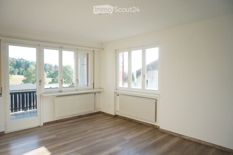 Empty room with wooden flooring, white walls, large windows, radiators, and a sliding door leading to a balcony.