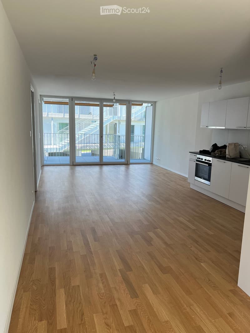 Empty room with wooden flooring, white walls, and a small kitchen area with cabinets and an oven. Large sliding glass doors lead to a balcony.