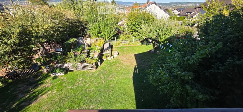 A well-maintained garden with a grassy lawn, surrounded by trees and shrubs. There is a wooden fence on one side and a small shed in the background. The garden is enclosed and features a variety of plants and flowers.