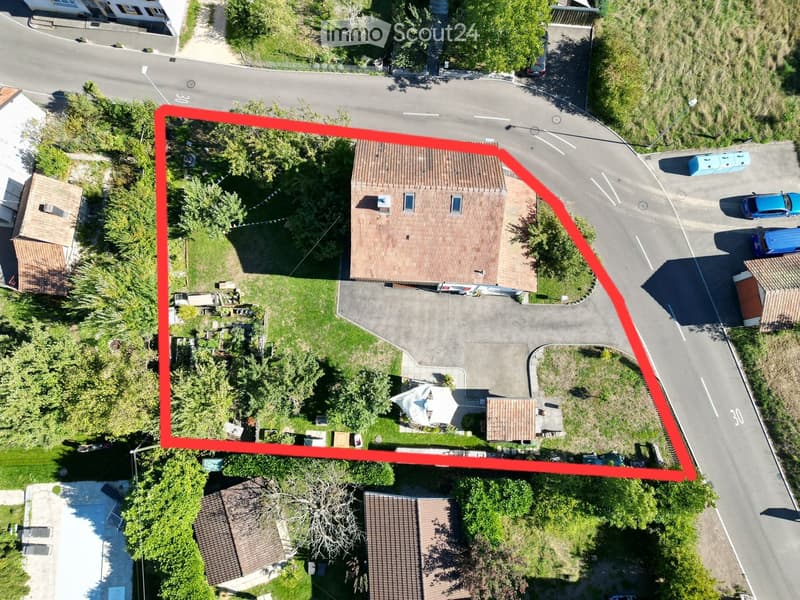 Aerial view of a two-story house with a tiled roof, surrounded by a driveway, lawn, and trees. The house has multiple windows and a small patio area with an umbrella.