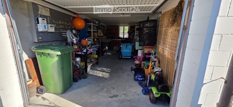 Garage with various stored items including a green trash bin, lawn mower, bicycles, children's toys, and storage shelves.