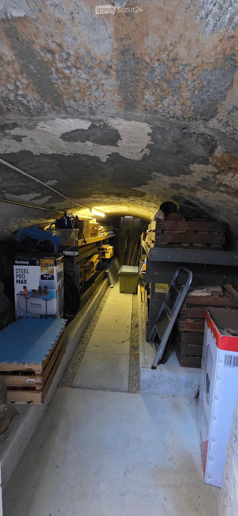 Underground corridor with exposed stone walls, concrete floor, and storage items including shelves, boxes, and a ladder.