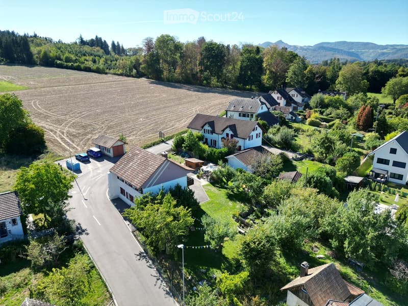 Aerial view of a residential area with multiple houses, surrounded by greenery and a road. The houses have varied architectural styles and are set in a rural landscape with fields and trees in the background.
