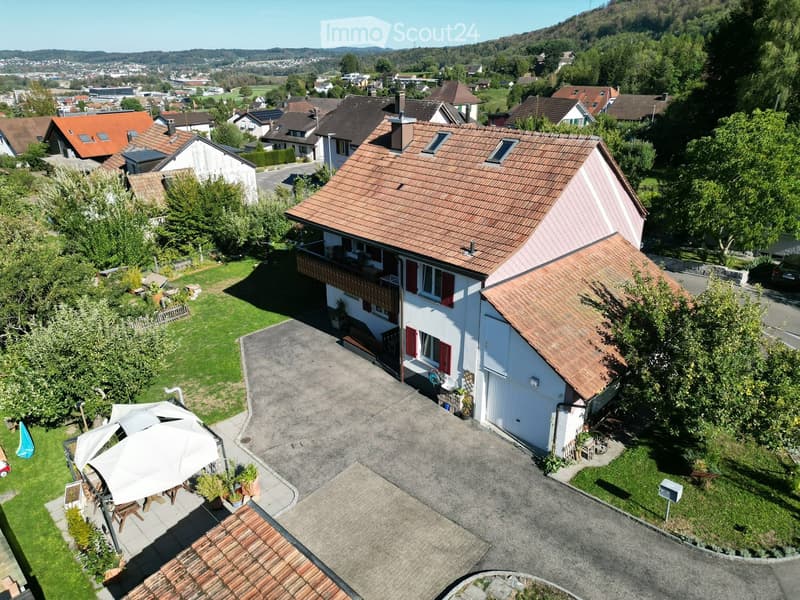 Aerial view of a two-story house with a red-tiled roof, white exterior, and a balcony. The property includes a driveway, a small garden area with outdoor furniture, and is surrounded by other houses and greenery.