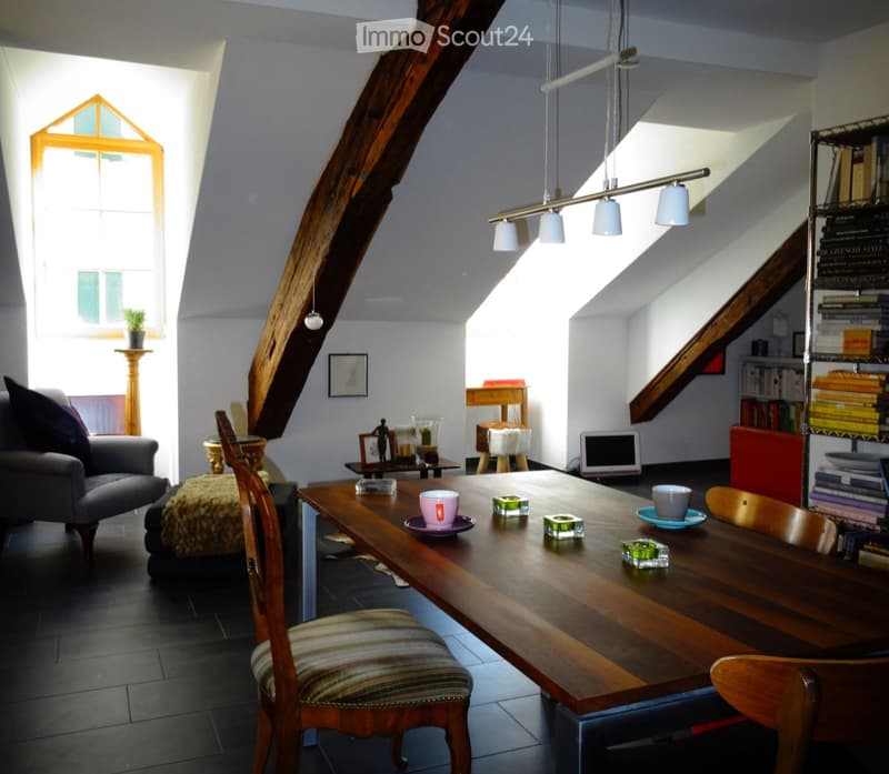 A cozy living room with a large wooden dining table, chairs, a sofa, a laptop, and bookshelves. The room features exposed wooden beams and ample natural light from large windows.