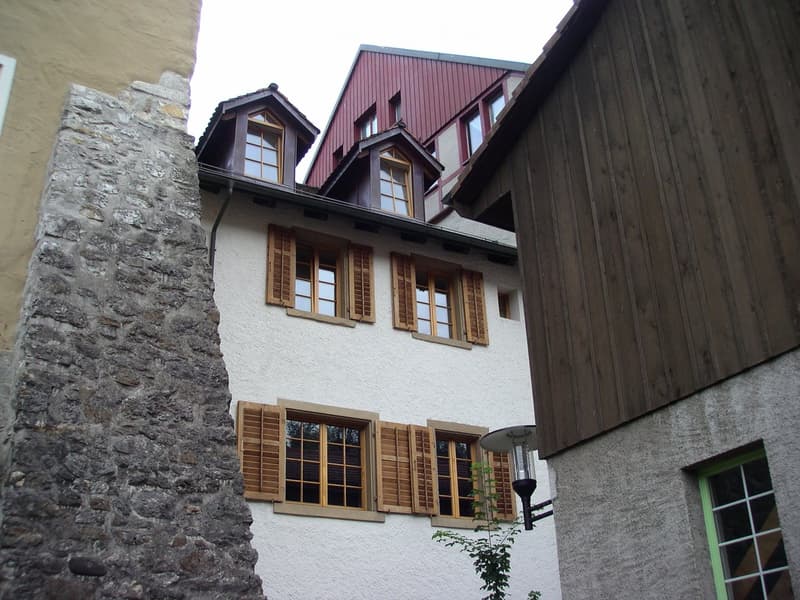A multi-story building with white walls, wooden shutters, and a red-tiled roof. The structure features multiple windows and a small dormer window on the roof.