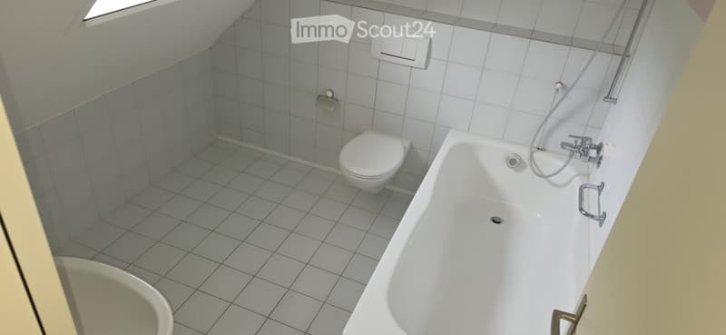 Modern bathroom with a bathtub, toilet, and tiled walls and floor. Features a shower hose attached to the wall and a window at the top left corner.