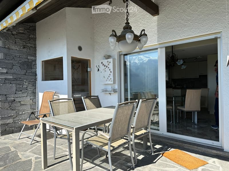 Outdoor patio with a glass table and several chairs, connected to a modern kitchen through a large sliding glass door. The patio features a stone wall and a hanging light fixture.