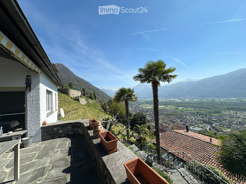 Balcony with stone railing, potted plants, and a panoramic view of a town and mountains.