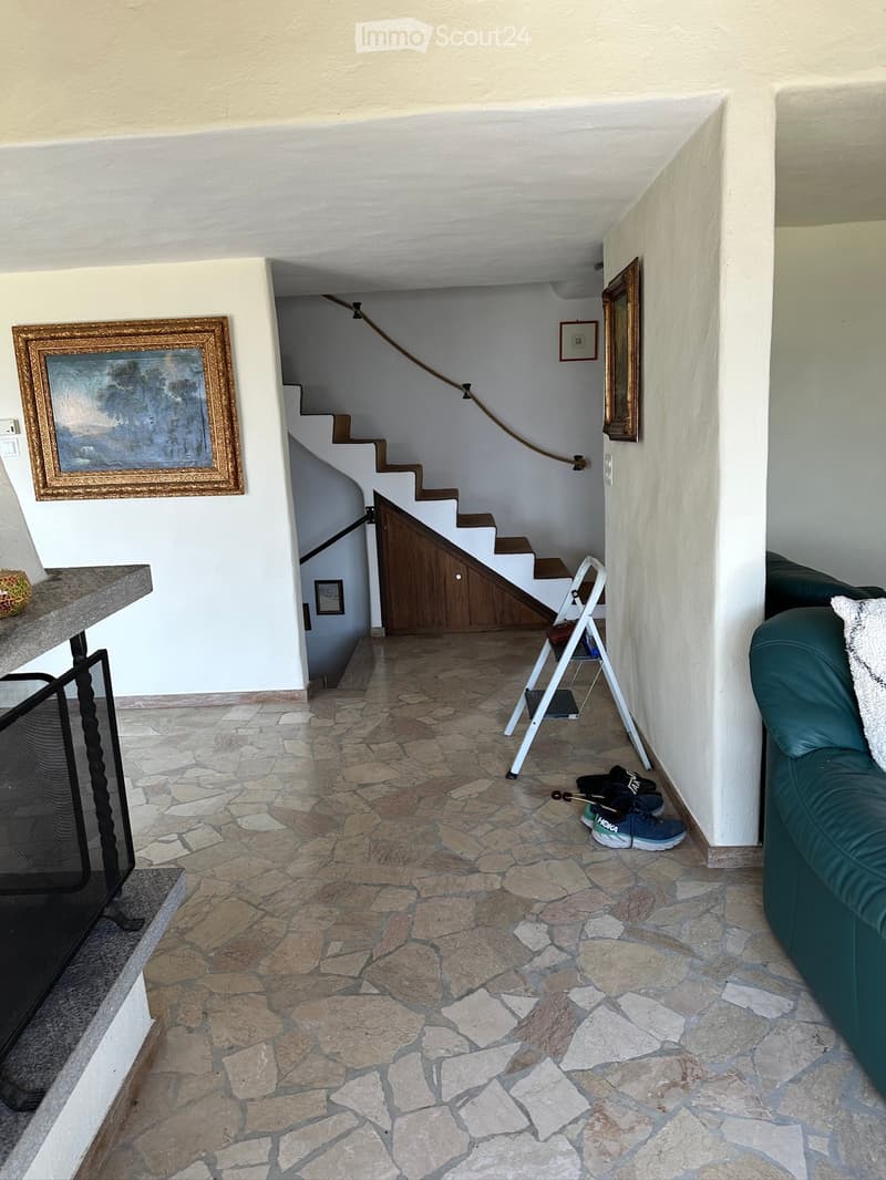 A hallway with a stone-tiled floor, a green leather sofa, a small white stool, a framed picture on the wall, a white folding stool ladder, and a pair of shoes on the floor.
