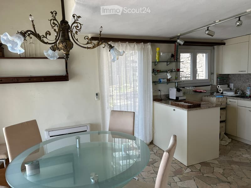 A modern, well-lit dining and kitchen area with a glass dining table, four chairs, white kitchen cabinets, a refrigerator, and a coffee maker. The space features a chandelier, shelves, and a window with curtains.