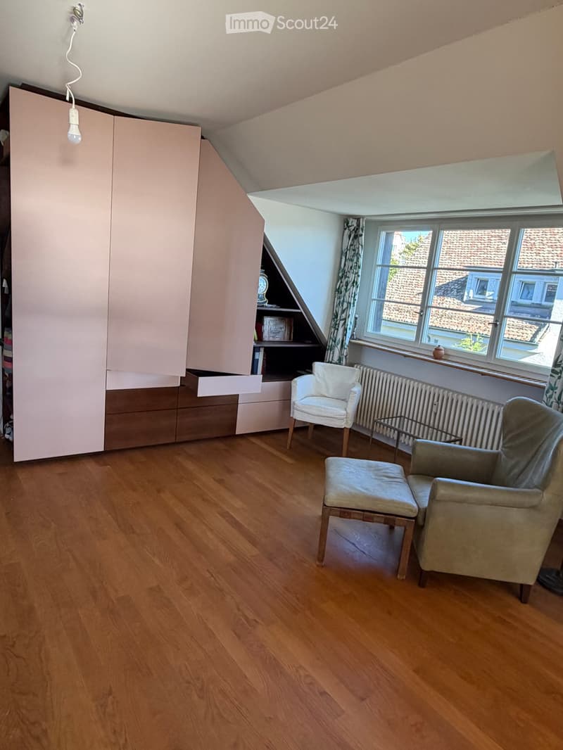 A cozy attic room with wooden flooring, large window, and modern furniture including a white armchair, a grey sofa, a small table, and a built-in cabinet with shelves.