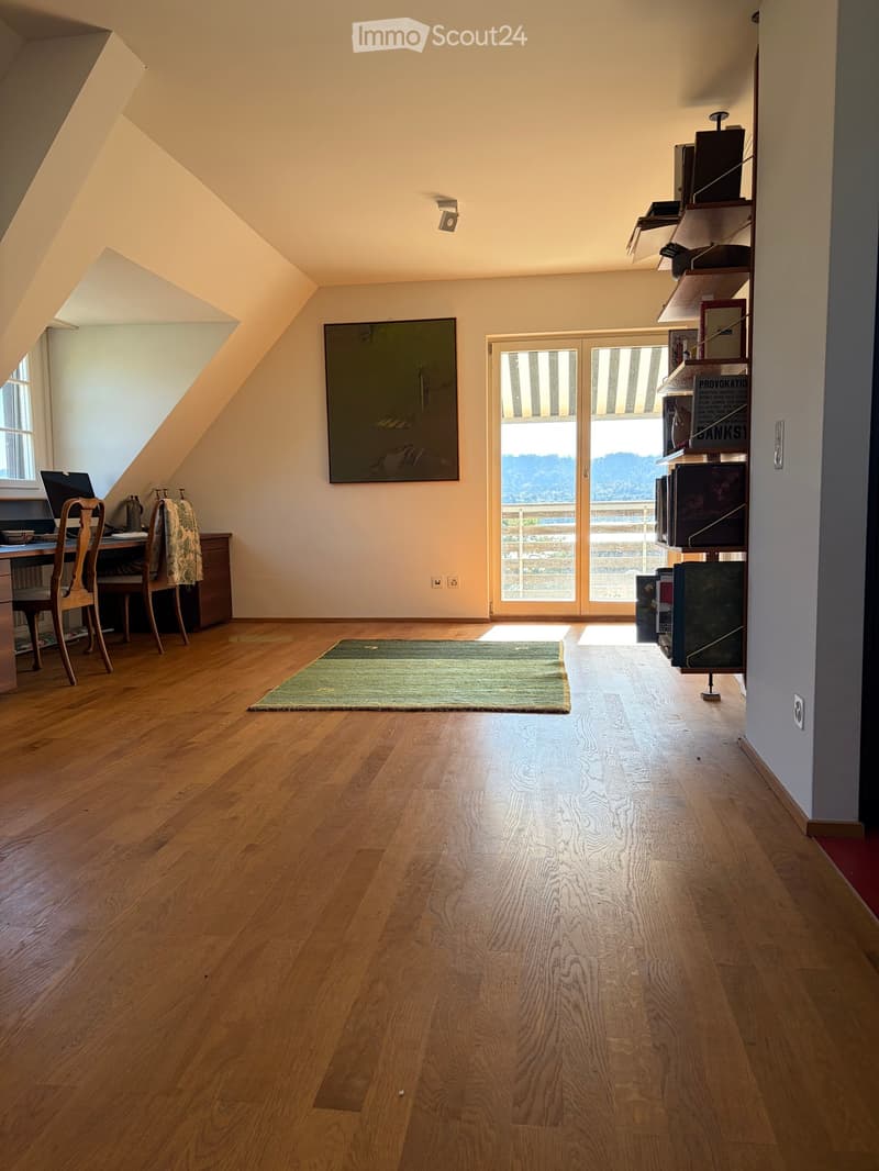 Bright, airy room with wooden flooring, large window, and a door leading to a balcony. Contains a desk with a chair, laptop, and a shelf with books and decorative items.