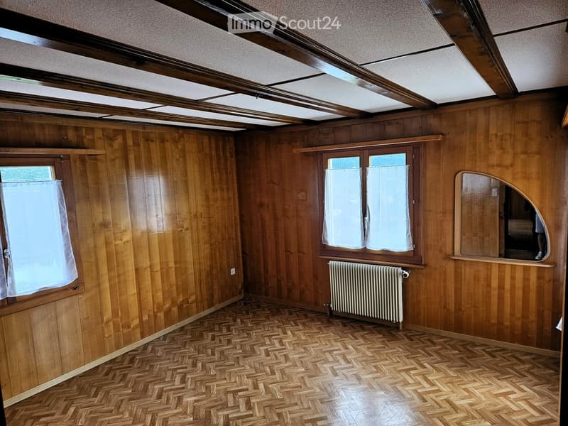 Empty room with wooden walls, herringbone wooden flooring, and large windows with white curtains. Features a radiator and a small arched mirror on the wall.