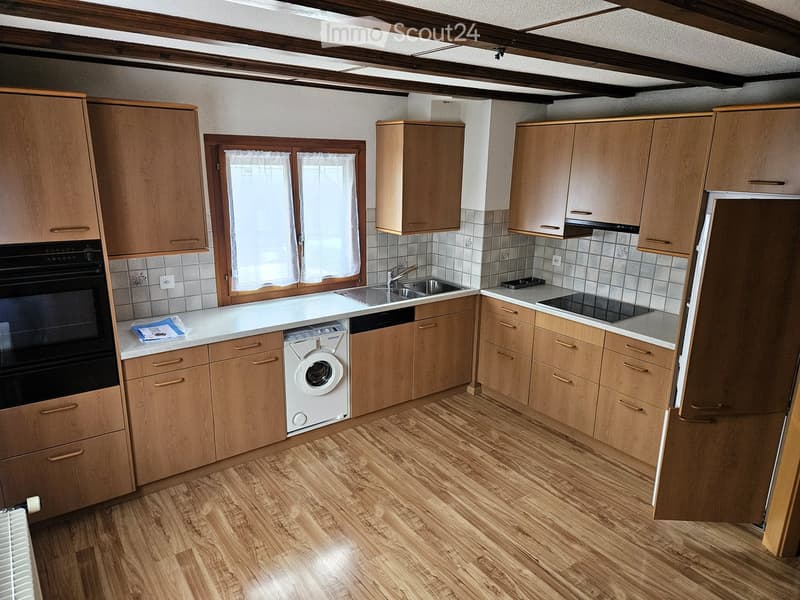 Bright kitchen with wooden cabinets, a white countertop, a sink, an oven, and a washing machine. The space features wooden flooring and a window with white curtains.