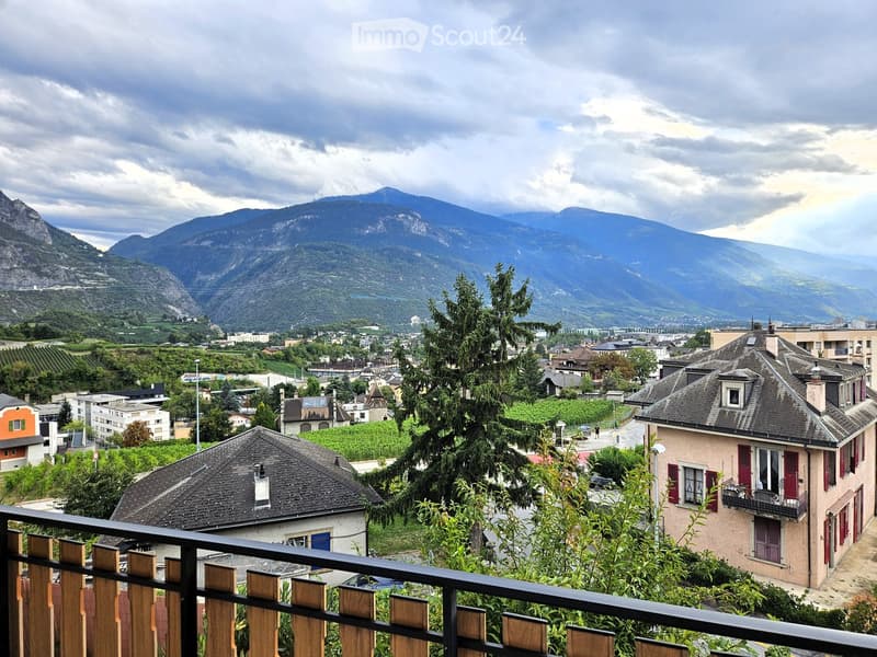 A scenic view from a balcony overlooking a town with houses, trees, and distant mountains under a partly cloudy sky.