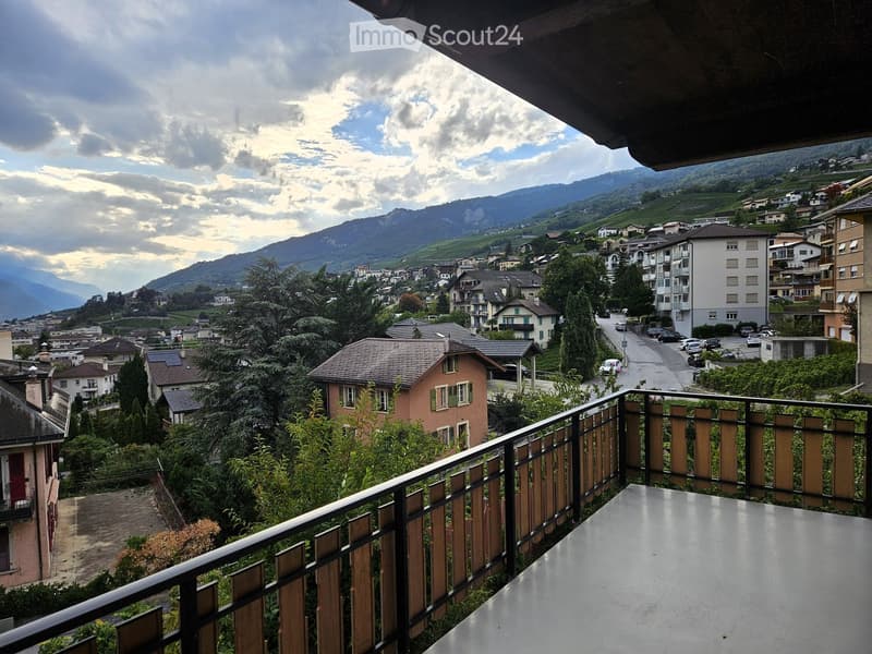 Balcony overlooking a town with houses, trees, and mountains in the background.