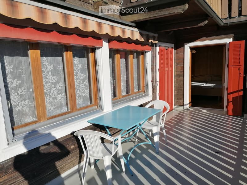 Balcony with a turquoise table and two white chairs, wooden decking, large windows with floral curtains, and a view into a bedroom with a bed.