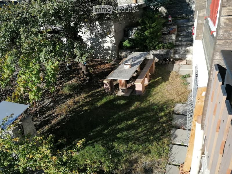 Garden area with a wooden bench, small table, and various plants, including a grapevine and shrubs. The space includes a stone wall, a small staircase, and a partially constructed wooden structure on the right side.