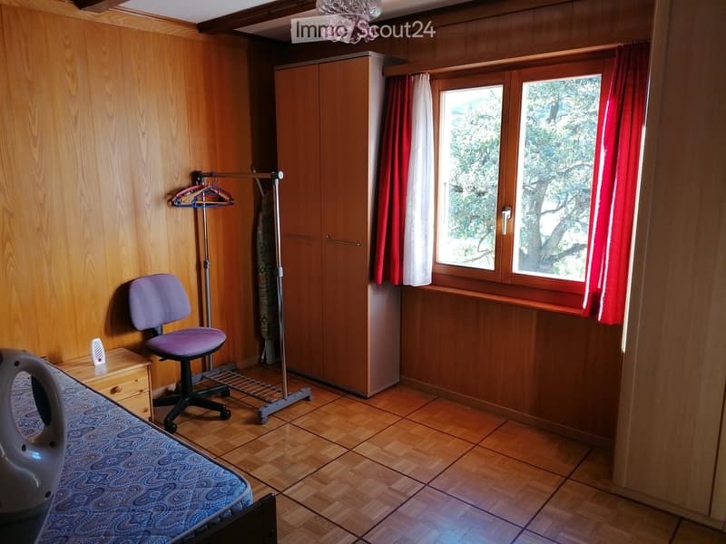 Cozy bedroom with wooden walls and floor, featuring a bed, a purple chair, a wardrobe, and a window with red curtains.