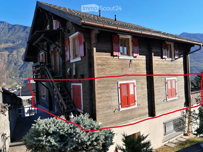 A two-story wooden house with red shutters, multiple windows, and a staircase leading to a balcony. The house is surrounded by greenery and situated in a mountainous area.