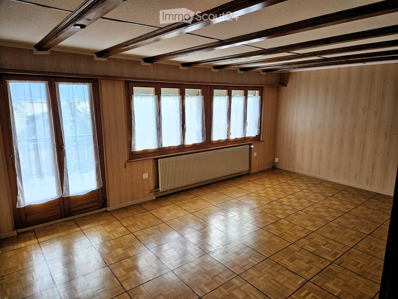 Empty room with wooden panel walls, wooden floor, large windows with white curtains, radiator, and exposed wooden ceiling beams.