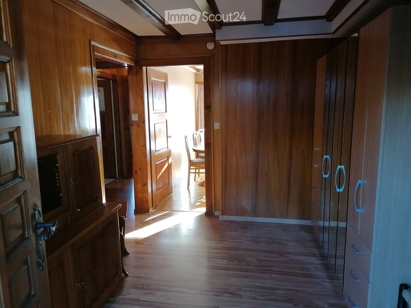 Wooden-paneled hallway with polished wooden floor, multiple doors, and a glimpse of a dining area with chairs and a table in the background.