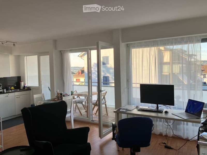A modern, well-lit room with a desk setup, a dining area, and a balcony view. The room features a wooden floor, large windows with sheer curtains, and a mix of contemporary and functional furniture.