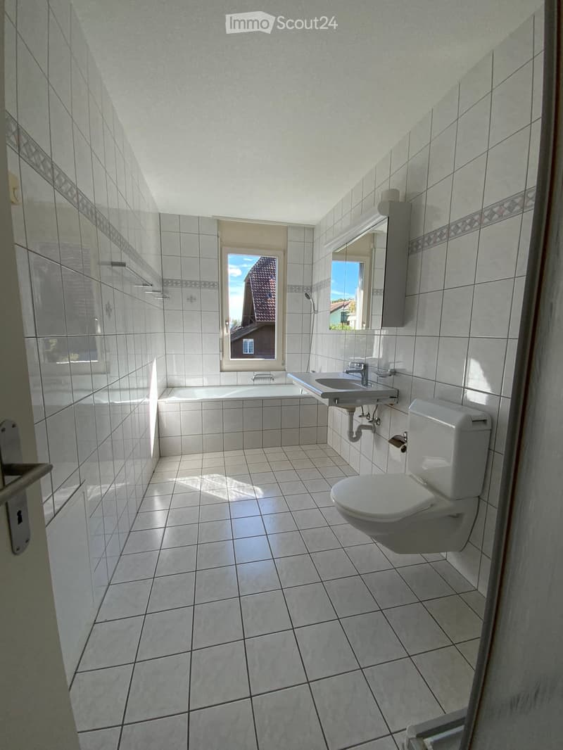 Bright bathroom with white tiled walls and floor, featuring a toilet, sink, bathtub, and a mirror above the sink. A window provides natural light.