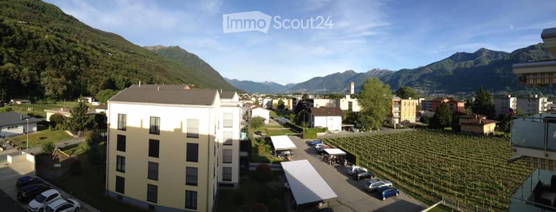 A multi-story residential building with a parking lot, surrounded by vineyards and a scenic mountain view.