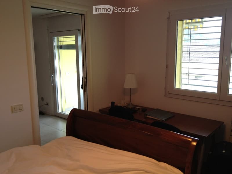 Bedroom with a wooden bed, desk, chair, lamp, and laptop. Features large windows and a sliding glass door leading to a balcony.