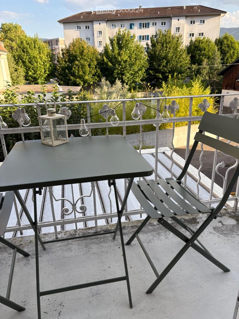 Balcony with a small table, two chairs, and a lantern, overlooking a cityscape with buildings and trees.