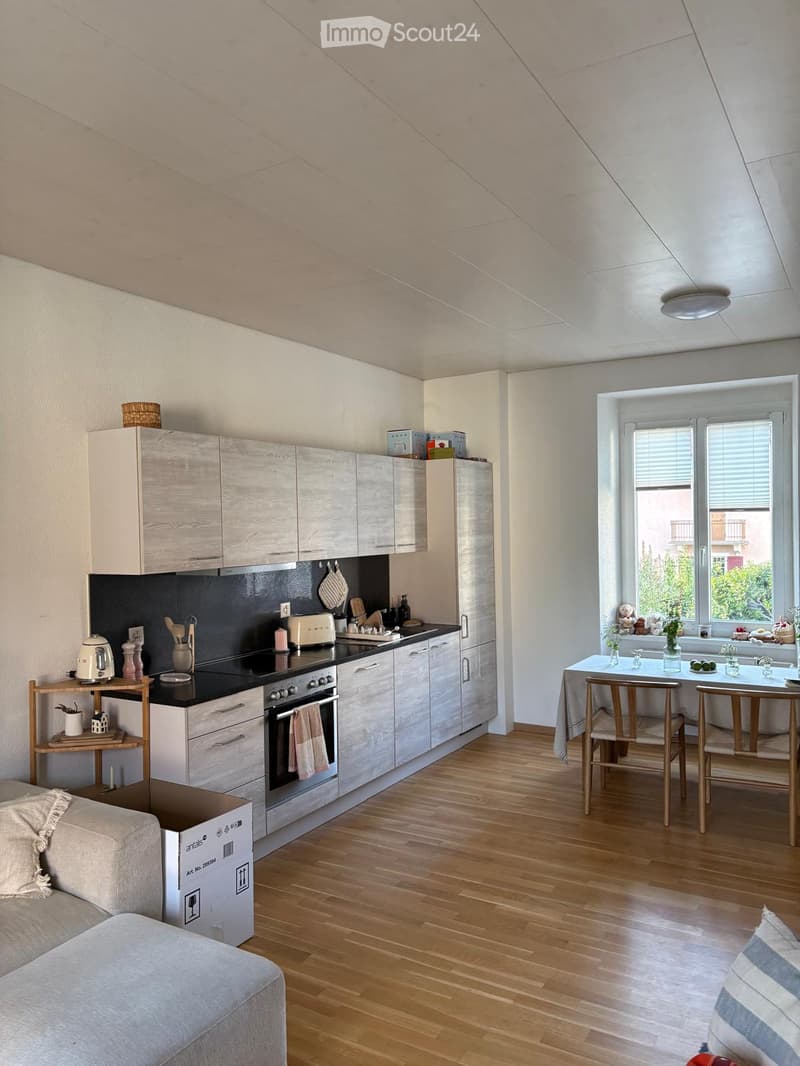 A modern, open-concept kitchen and living area with light wood flooring, white cabinets, and a black countertop. The kitchen features an oven, refrigerator, and various appliances. A small dining table with chairs is set near a large window with blinds, a