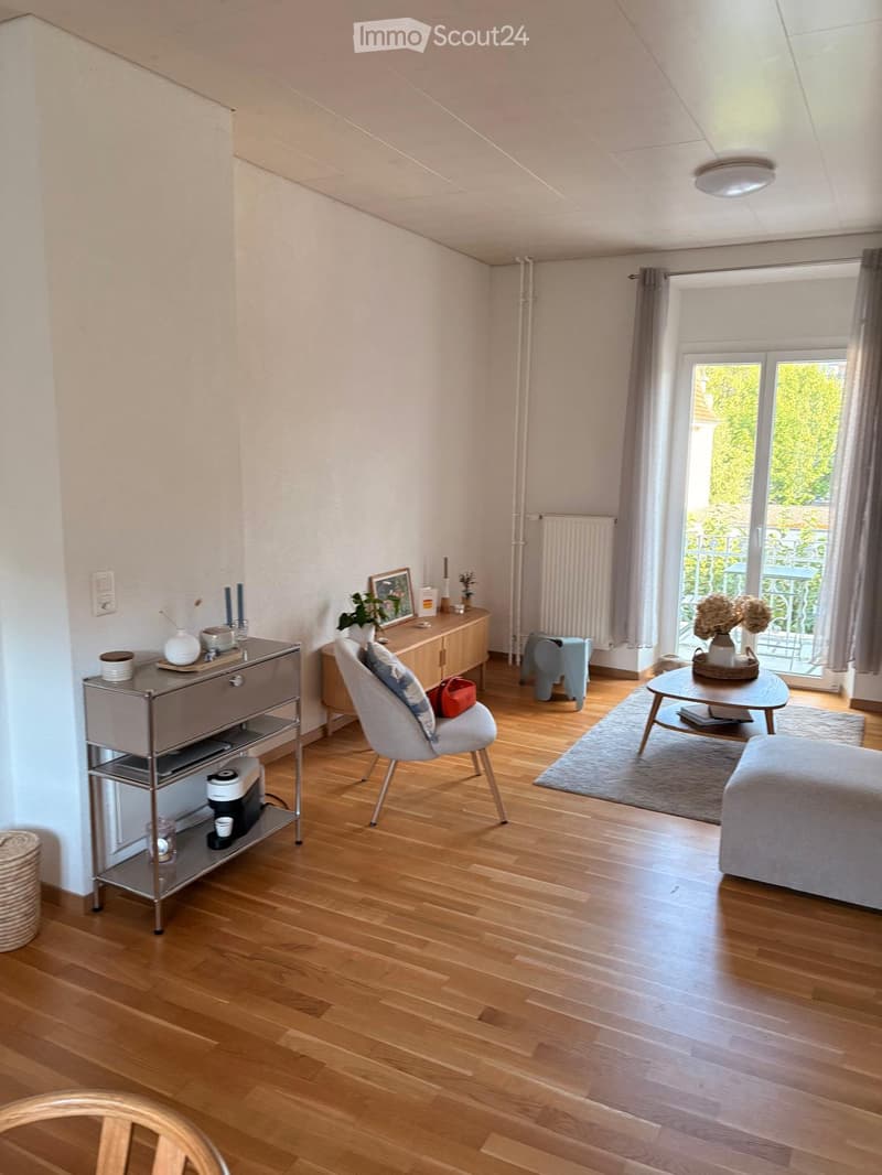 Bright living room with wooden flooring, large window with balcony access, modern furniture including a sofa, chair, coffee table, and side cabinet.