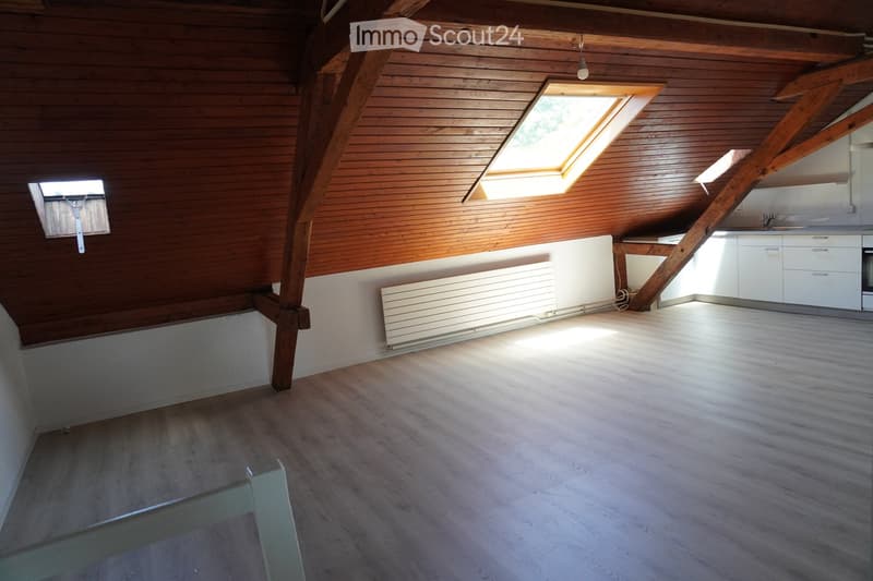 An empty attic space with wooden beams, skylights, and a small kitchen area in the corner. The floor is wooden, and the walls are painted white with wooden paneling on one side.