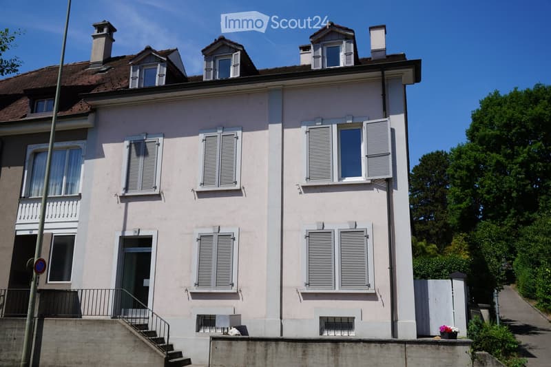 Two-story residential house with light pink facade, multiple windows with shutters, and a small balcony on the first floor.