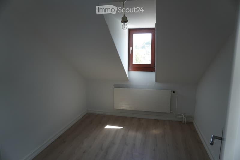Empty attic room with wooden flooring, white walls, a single window with wooden frame, and a radiator. No furniture or personal items present.