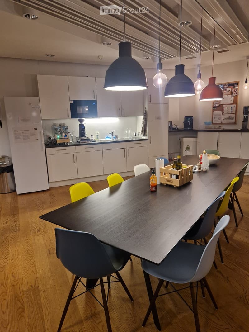 Modern kitchen with white cabinets, a long wooden dining table surrounded by colorful chairs, and various appliances including a refrigerator, microwave, and coffee machine.