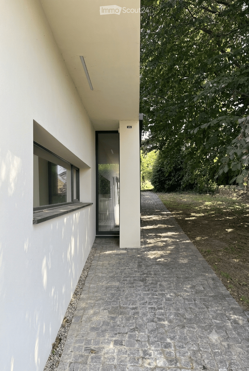 A paved pathway leads to a modern building entrance with large windows and a sleek design, surrounded by greenery.