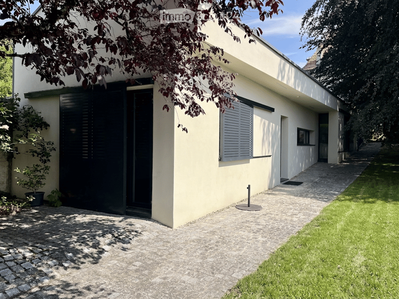 Modern single-story house with light yellow walls, black shutters, and a paved entrance path. A small potted plant is near the entrance, and the house is partially shaded by tree branches.