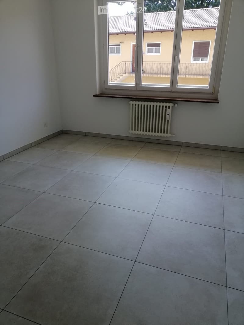 Empty room with tiled floor, white walls, radiator, and large window with view of a building and balcony.