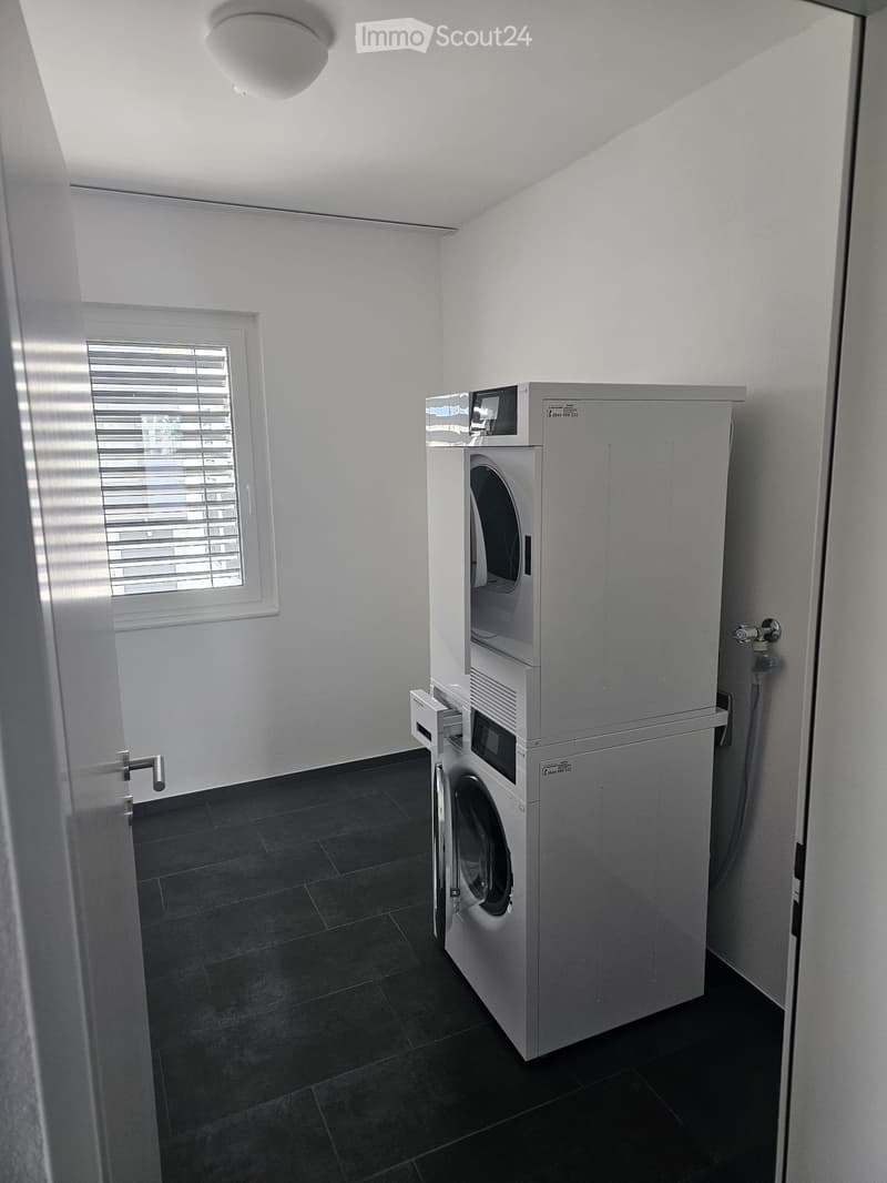 A small, well-lit room with a washing machine and a window with blinds.