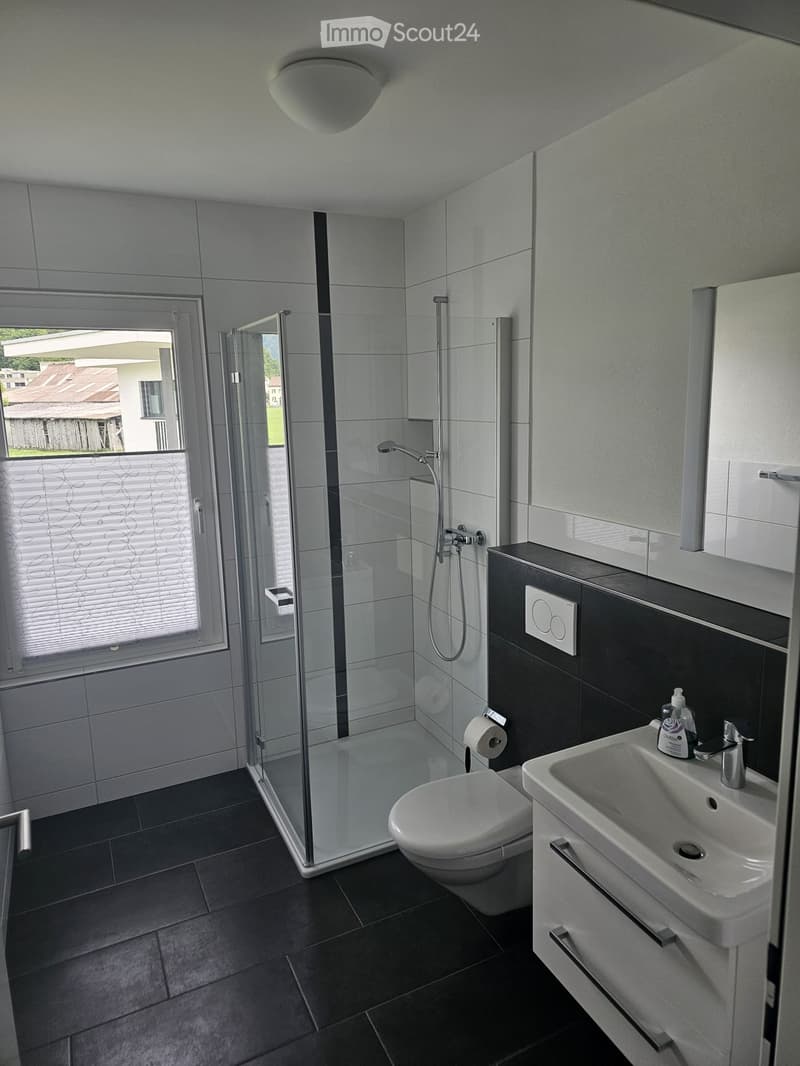 Modern bathroom with dark tiled floor, white toilet, sink with faucet, glass-enclosed shower, and a window with blinds.