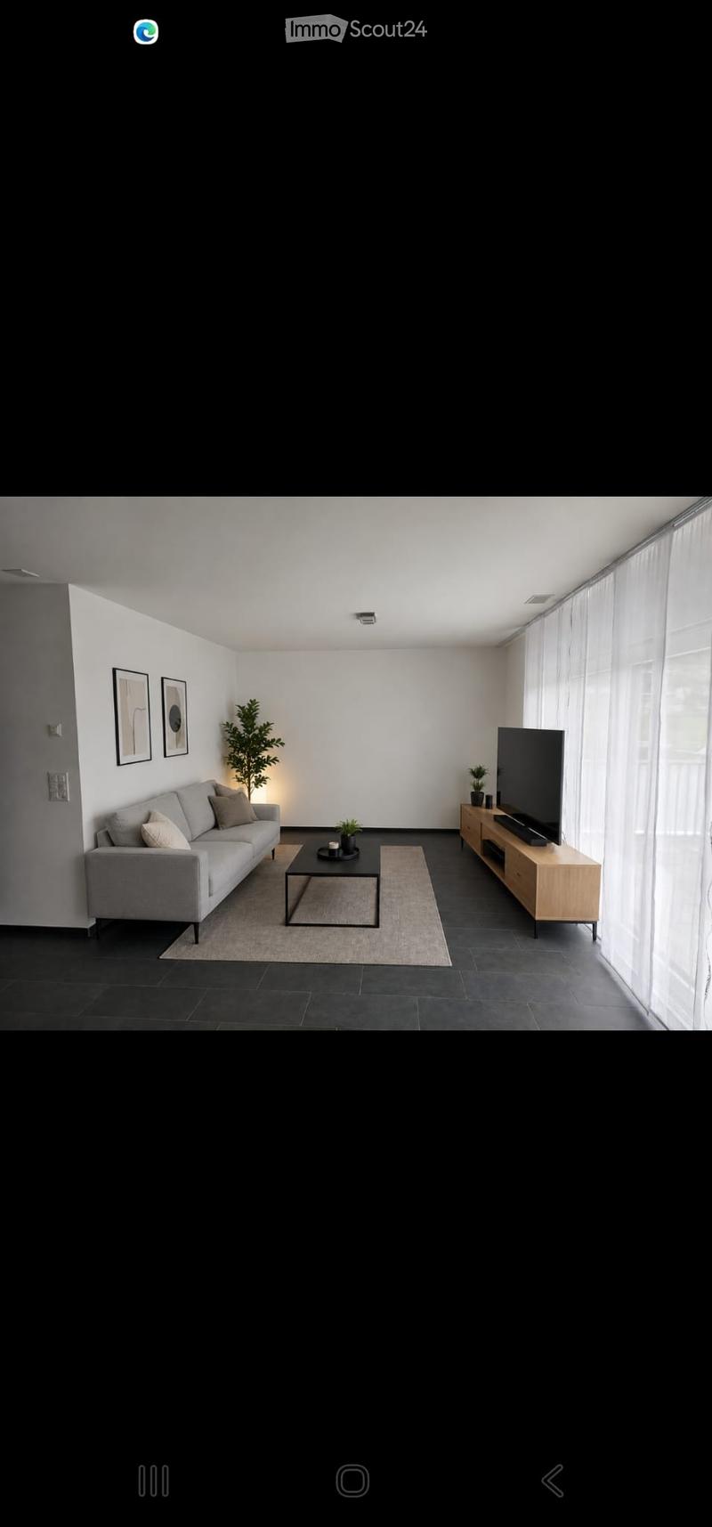 Modern living room with a grey sofa, coffee table, TV stand, and decorative plants. Large windows with sheer curtains allow natural light to fill the space.