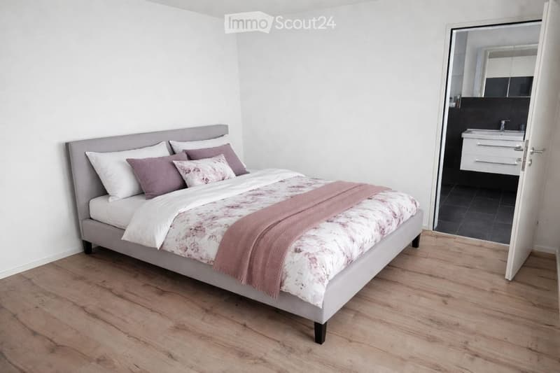 Modern bedroom with a grey upholstered bed, white and pink bedding, and a pink throw. The room features light wooden flooring and an open door leading to a small bathroom with a sink and mirror.