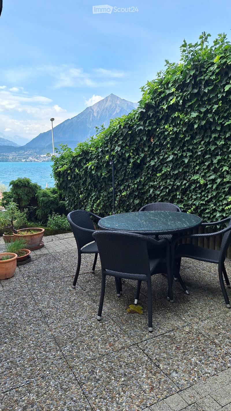 Outdoor patio with a glass table and four black wicker chairs, surrounded by potted plants and a lush green hedge, overlooking a scenic lake and mountain view.
