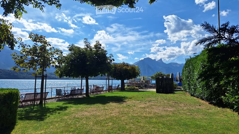 A serene garden area with a grassy lawn, benches, tables, and chairs, overlooking a lake with mountains in the background.