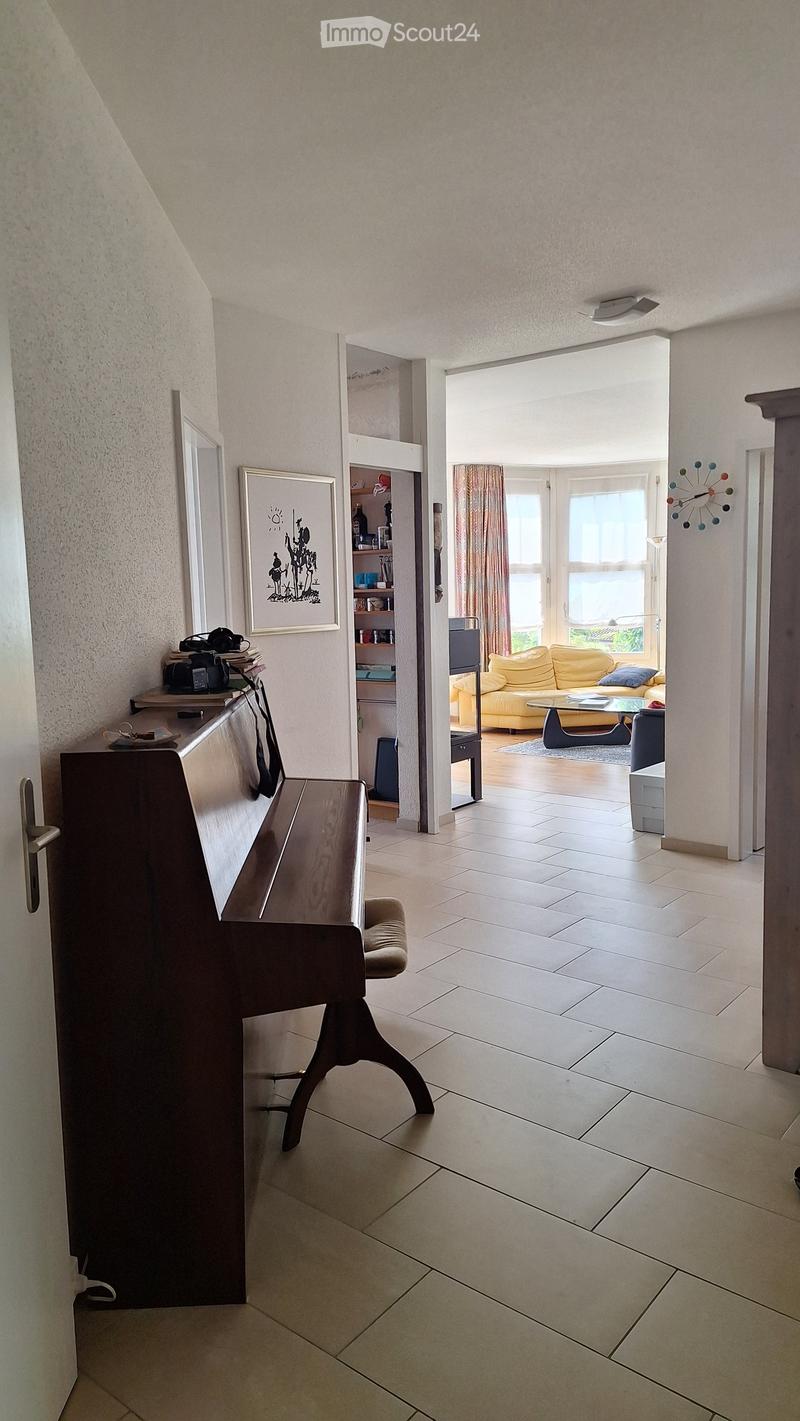 Bright hallway with tiled floor, open doorway leading to a living room with a yellow sofa, glass coffee table, and large windows. A wooden bench and shelves are visible in the hallway.